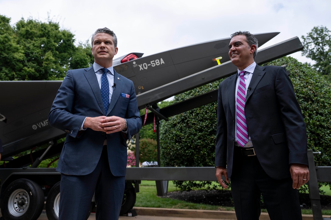 Defense Secretary Pete Hegseth and another civilian stand and talk in front of an unmanned aerial vehicle on display outdoors.