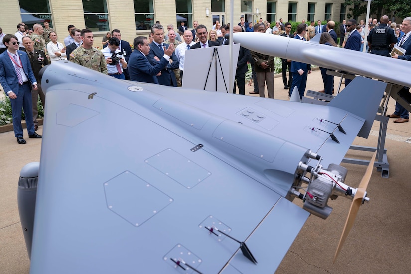 A crowd of military and civilian personnel stand around a drone on display.