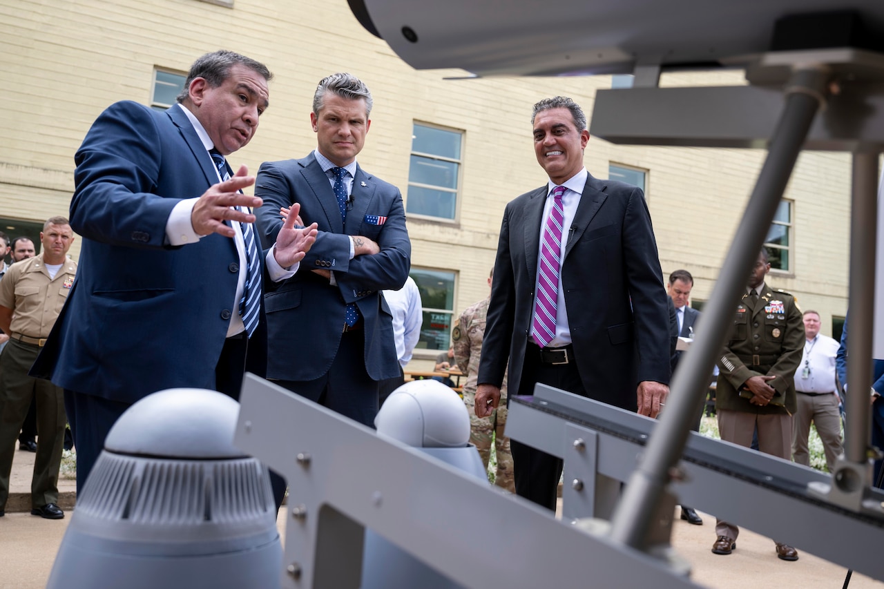 A man points out details of a drone on display to two other men. There is a crowd of onlookers behind them.