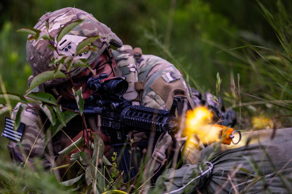 A soldier in combat gear and lying behind sandbags in a grassy area fires a rifle.