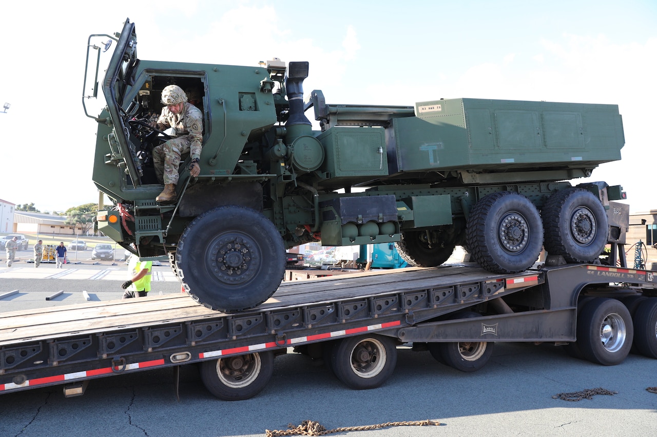 A large military vehicle is driven onto a flatbed trailer. A soldier in the driver's seat has the door open and is looking out.