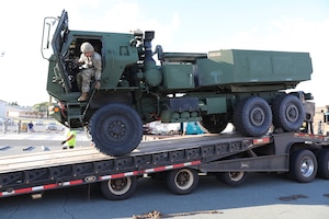 A large military vehicle is driven onto a flatbed trailer. A soldier in the driver's seat has the door open and is looking out.