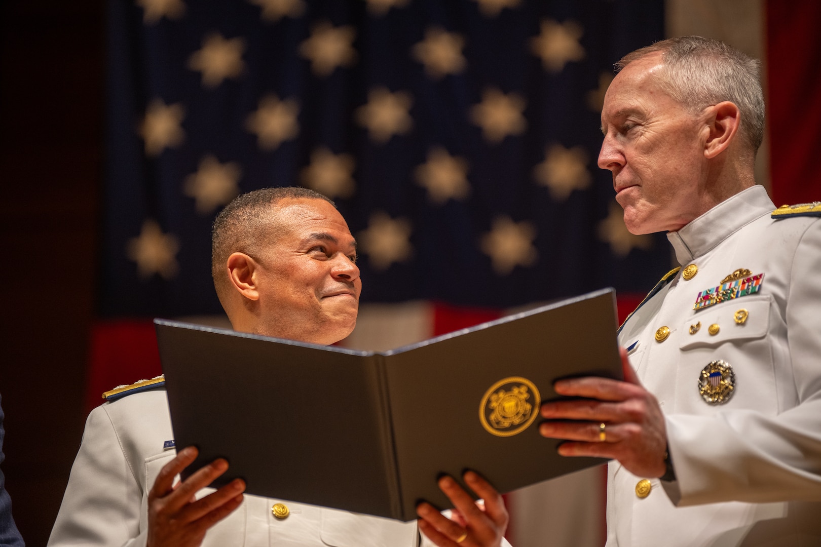 U.S. Coast Guard Rear Adm. Michael Johnston (left) smiles at Acting Commandant of the Coast Guard Adm. Kevin Lunday while receiving an award during the change of command ceremony inside Leamy Hall at the U.S. Coast Guard Academy (USCGA), New London, Connecticut, July 16, 2025. Johnston was relieved by Rear Adm. Gregory Rothrock who officially became the 44th superintendent of USCGA. (U.S. Coast Guard photo by Petty Officer Third Class Leo Avila.)