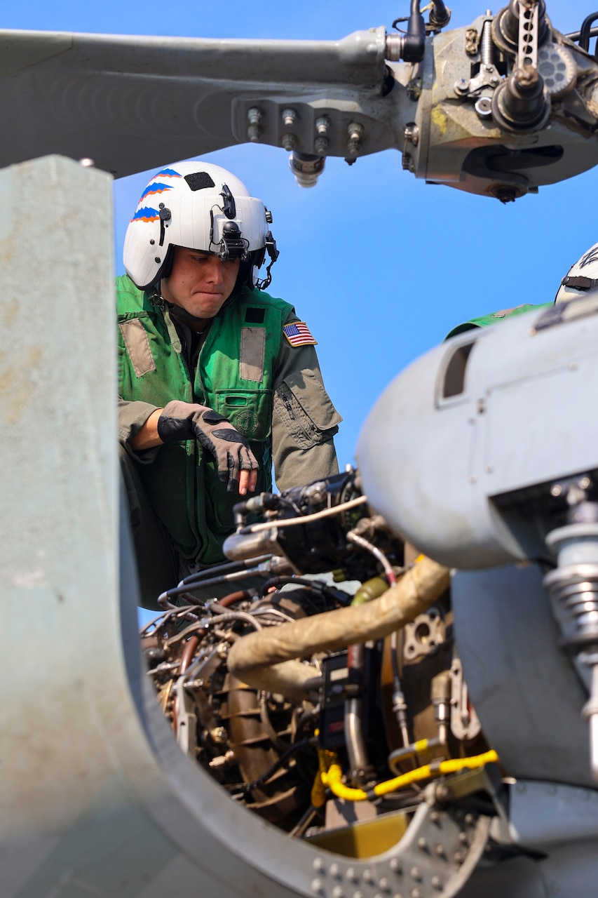 A sailor wearing a flight helmet checks parts on a helicopter under a blue sky.