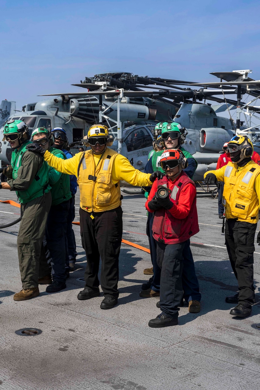 Standing sailors and Marines hold hoses on the deck of a ship, with stationary helicopters in the background.