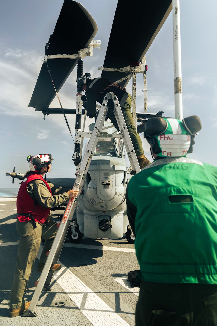 Two Marines, one preparing to ascend a ladder, work to secure a helicopter to the deck of a ship under a gray sky.