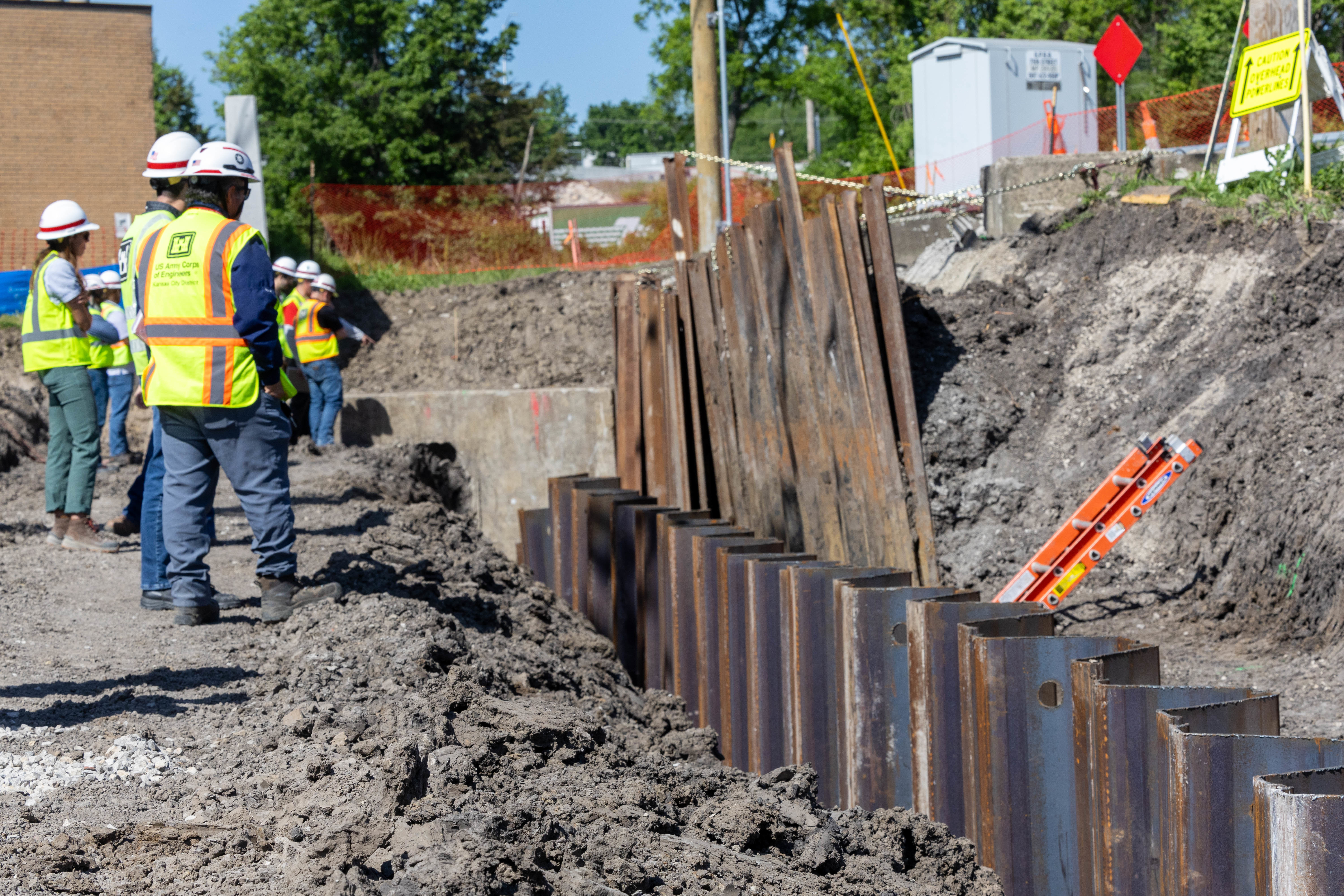 People in yellow safety vests and hardhats stand on dirt with an orange safety fence in the background and brown metal sheets to the right.