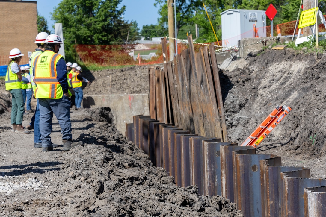 People in yellow safety vests and hardhats stand on dirt with an orange safety fence in the background and brown metal sheets to the right.