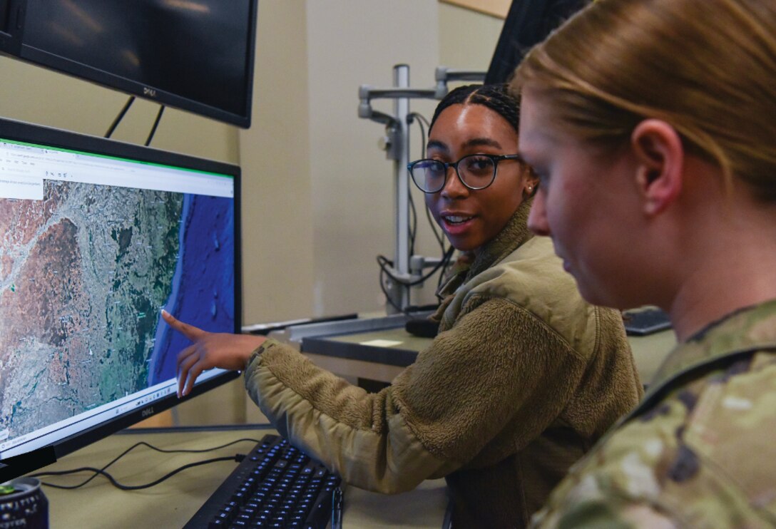 Two military personnel analyze satellite imagery on a computer monitor. One woman, wearing glasses and a fleece jacket, points to a section of the map displayed on the screen, while the other, in camouflage uniform, observes attentively. The setting appears to be a military or intelligence operations room equipped with computers and monitors.
