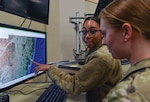 Two military personnel analyze satellite imagery on a computer monitor. One woman, wearing glasses and a fleece jacket, points to a section of the map displayed on the screen, while the other, in camouflage uniform, observes attentively. The setting appears to be a military or intelligence operations room equipped with computers and monitors.