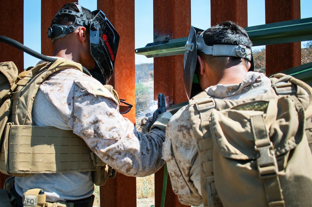 Two Marines wearing face protection weld metal rods to an outdoor  security barrier on a sunny day.
