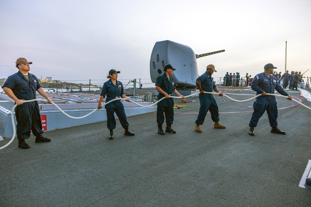 Five sailors hold a large, white rope on the deck of a ship, with a large gun and other sailors standing in the background.