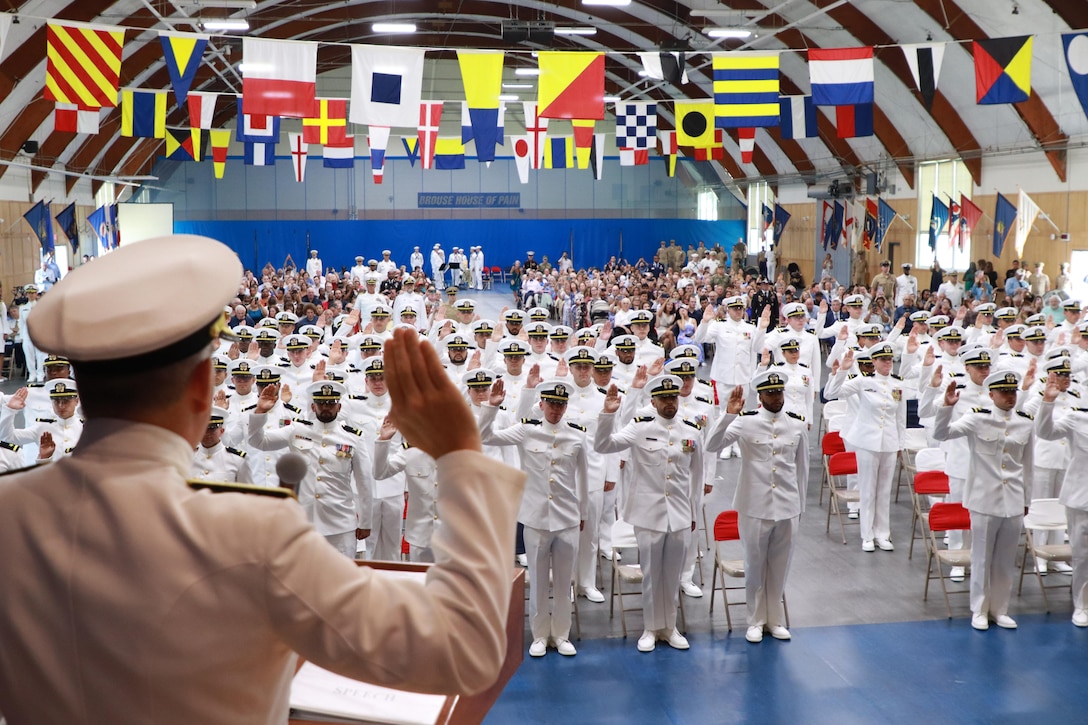 A sailor faces a group of sailors holding their right hands up in an auditorium with colorful flags hanging from the rafters.