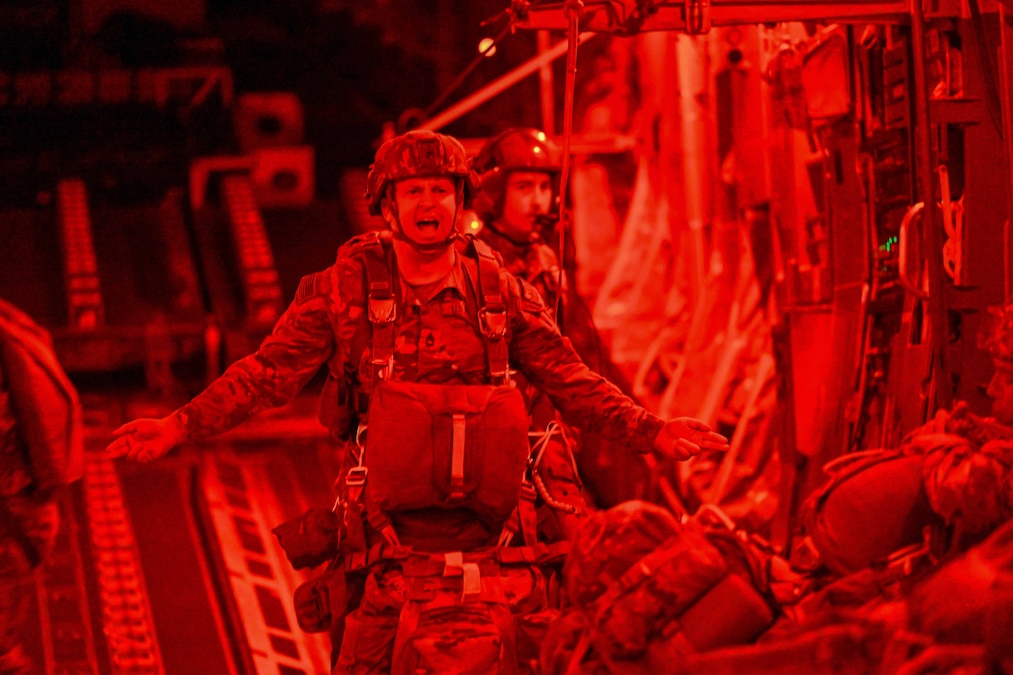 A soldier in parachute gear speaks to other soldiers inside a plane, surrounded by red light.