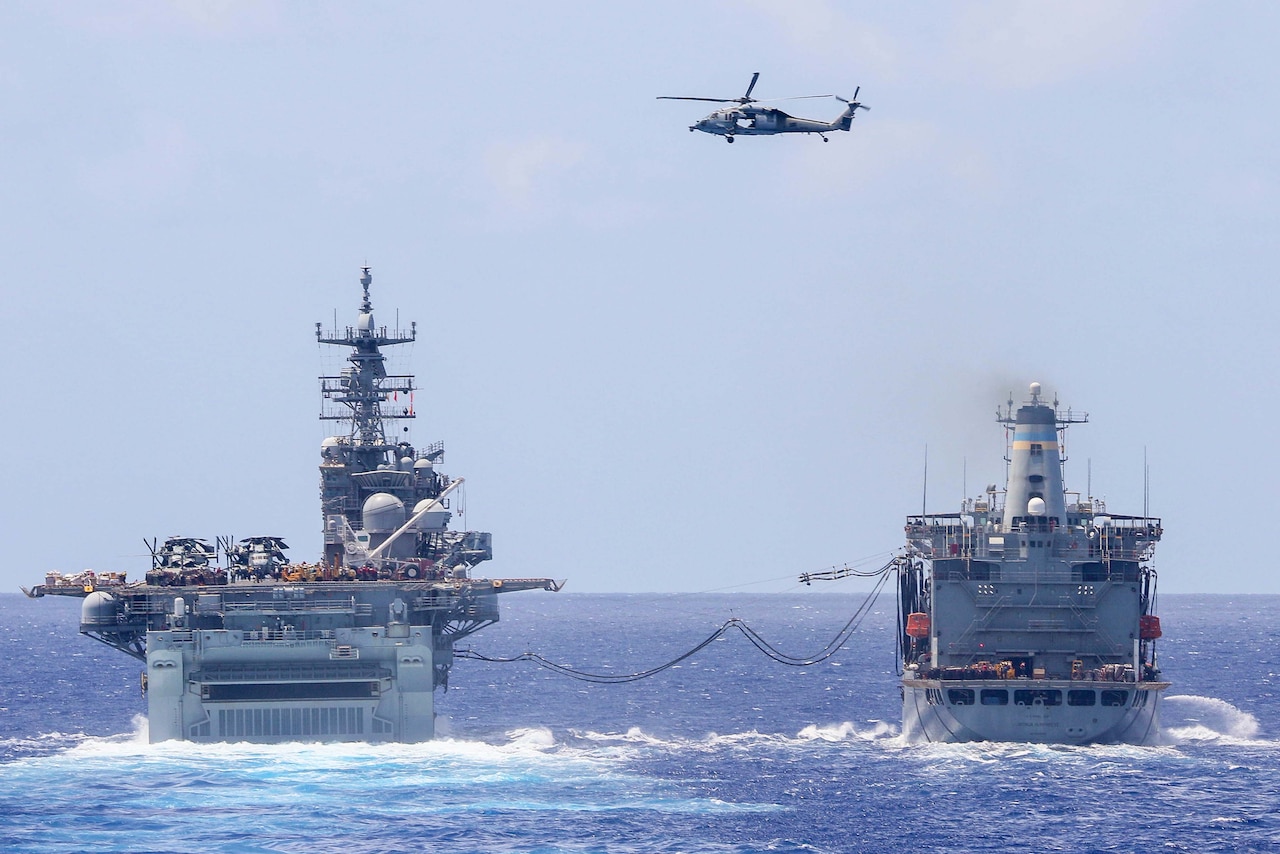 Two ships cruise near each other in the ocean, with large cables connecting the ships; a helicopter flies overhead.
