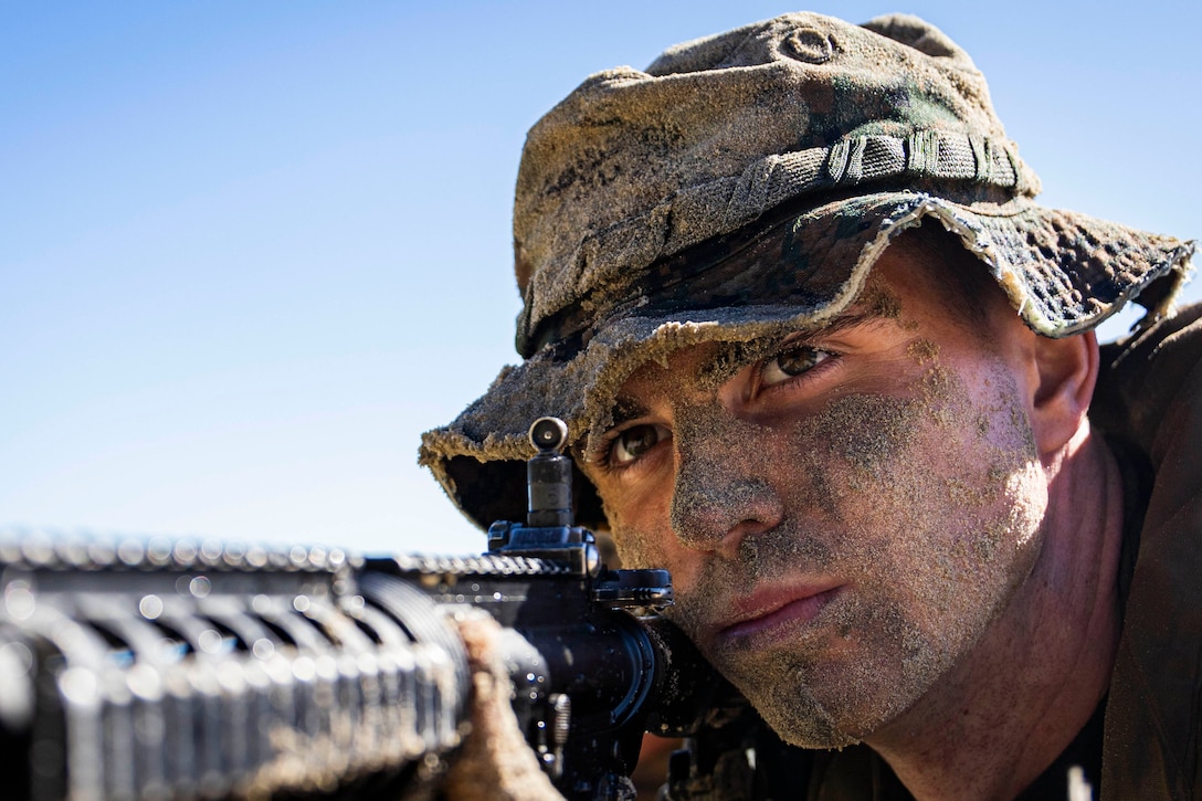A Marine in a hat aims a service weapon under blue skies, as seen from close-up.
