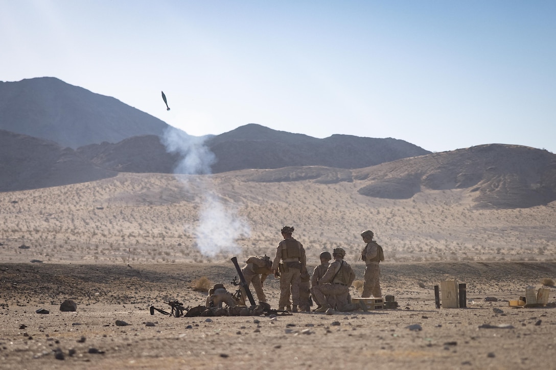 Marines fire a mortar system weapon in a desert setting under blue skies with mountains in the distance.