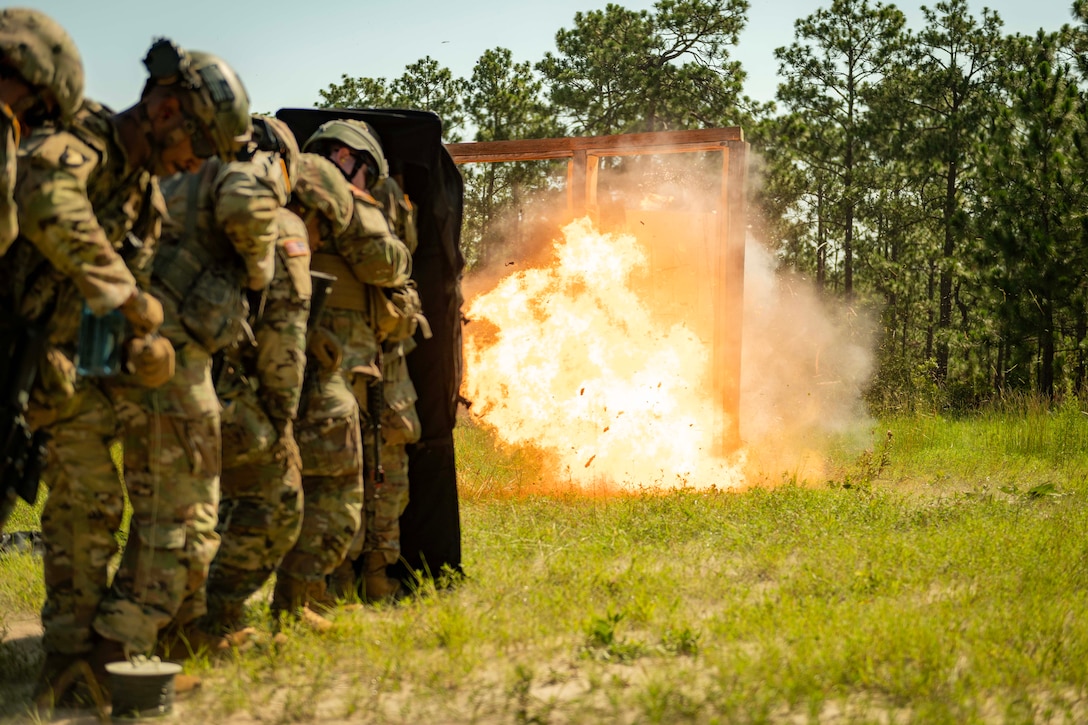 National Guard soldiers stand behind a shield as a wooden frame ignites into flame in a field.