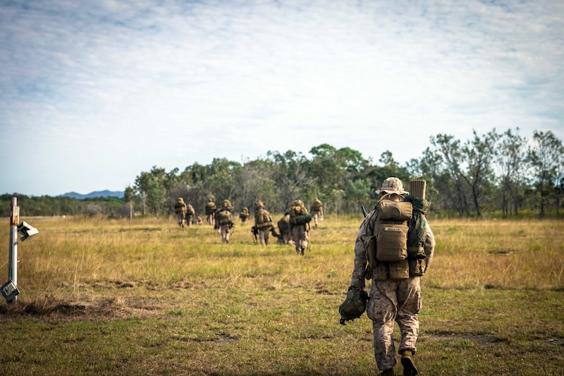 A group of Marines walk toward a tree line in the distance during the day.