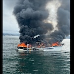 A boat is seen on fire and billowing smoke on the water.