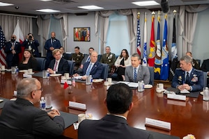 A group of people in business suits and dress military uniforms are gathered around a large wooden table.