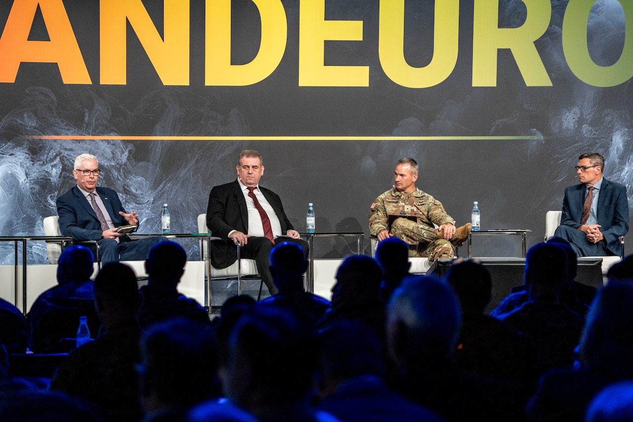 Three people dressed in business suits flank a soldier dressed in a military uniform. They are seated on a stage with a sign above their heads that reads LANDEURO.