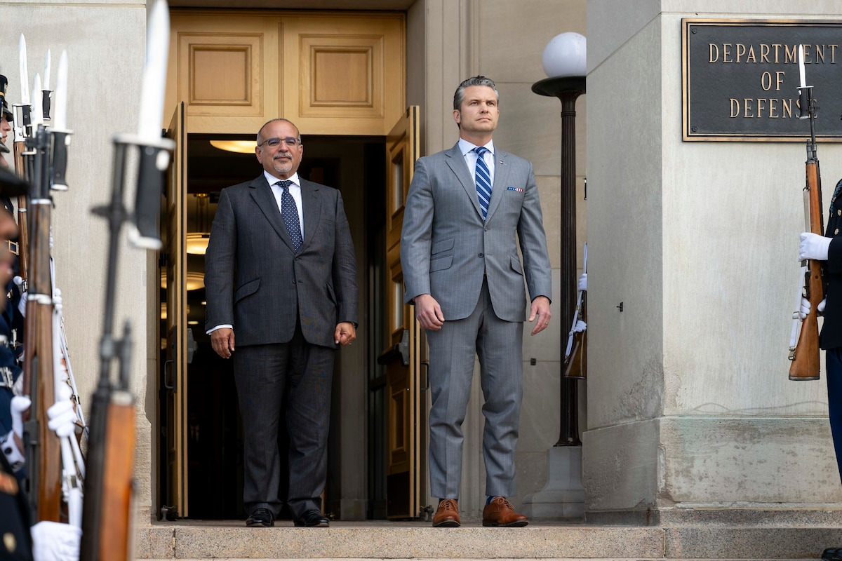 Two men in business suits are standing outside a building facing forward. A sign that reads “Department of Defense” is on a pillar off to the right. There is an open office building door behind the men.