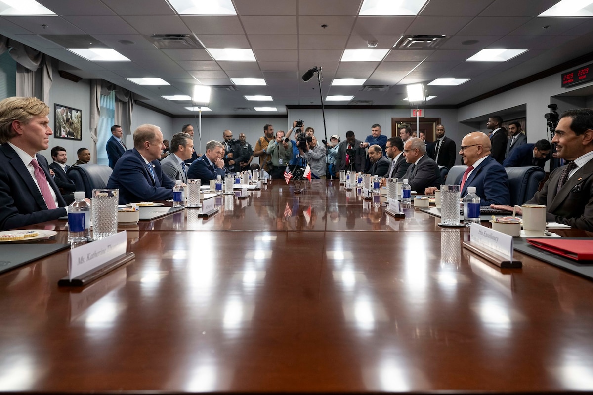 Men in business suits are seated around a large wooden table conversing from opposite sides. In the background, people with cameras and light kits are trying to capture photos of the meeting.