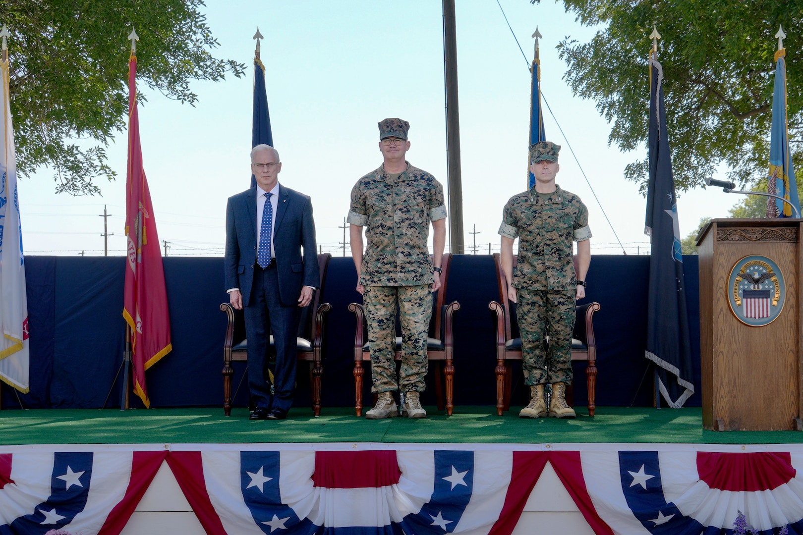 Two men wearing camouflage military uniforms and a man in a blue suit stand on a stage outdoors.