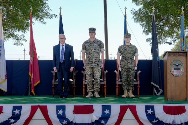 Two men wearing camouflage military uniforms and a man in a blue suit stand on a stage outdoors.