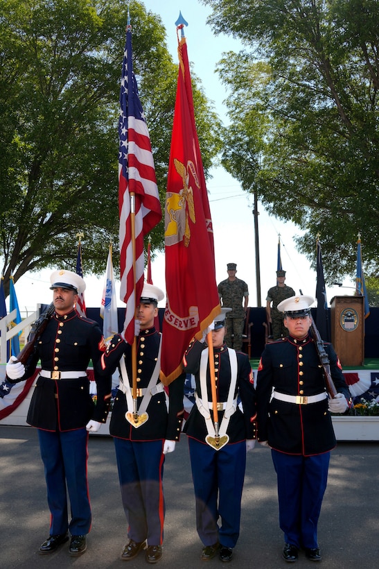 Four men wearing U.S. Marine Corps uniforms stand in front of a stage presenting the United States and U.S. Marine Corps flags.