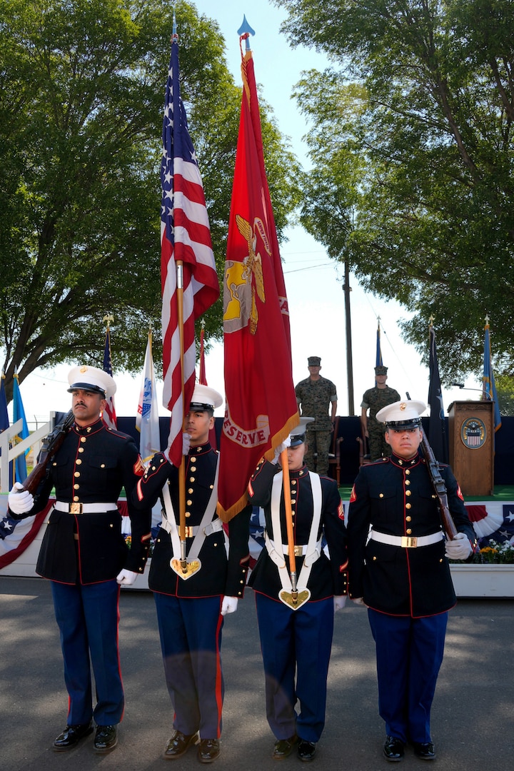 Four men wearing U.S. Marine Corps uniforms stand in front of a stage presenting the United States and U.S. Marine Corps flags.