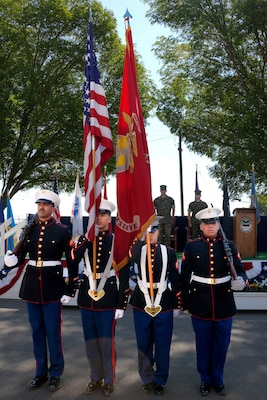 Four men wearing U.S. Marine Corps uniforms stand in front of a stage presenting the United States and U.S. Marine Corps flags.