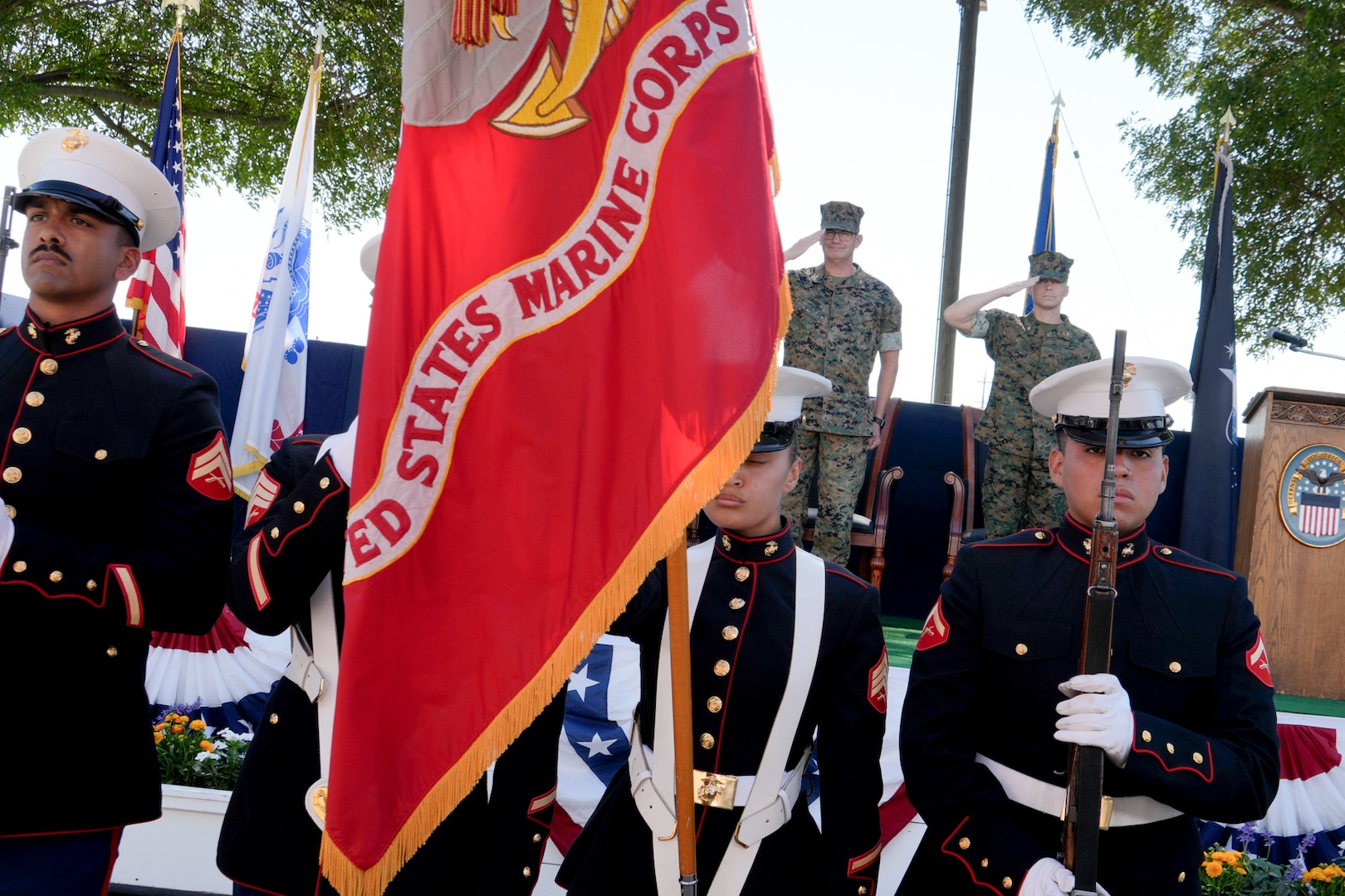 Four men in U.S. Marine Corps uniforms stand in front of a stage with the U.S. Marine Corps flag in front of them.