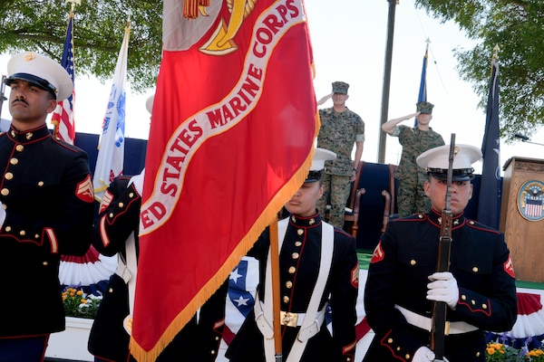 Four men in U.S. Marine Corps uniforms stand in front of a stage with the U.S. Marine Corps flag in front of them.