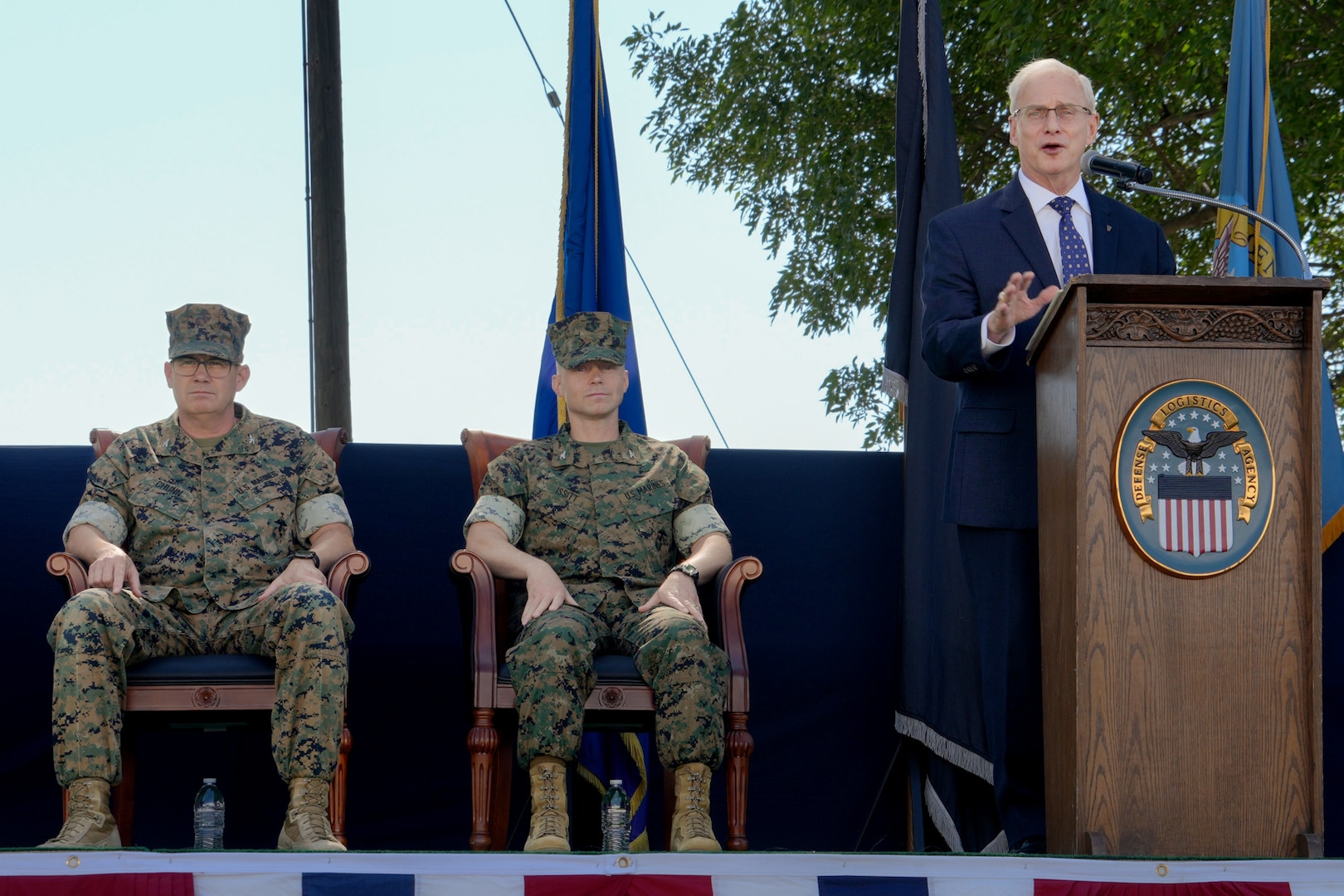 A man in a blue suit stands on a stage outdoors behind a lectern giving a speech.