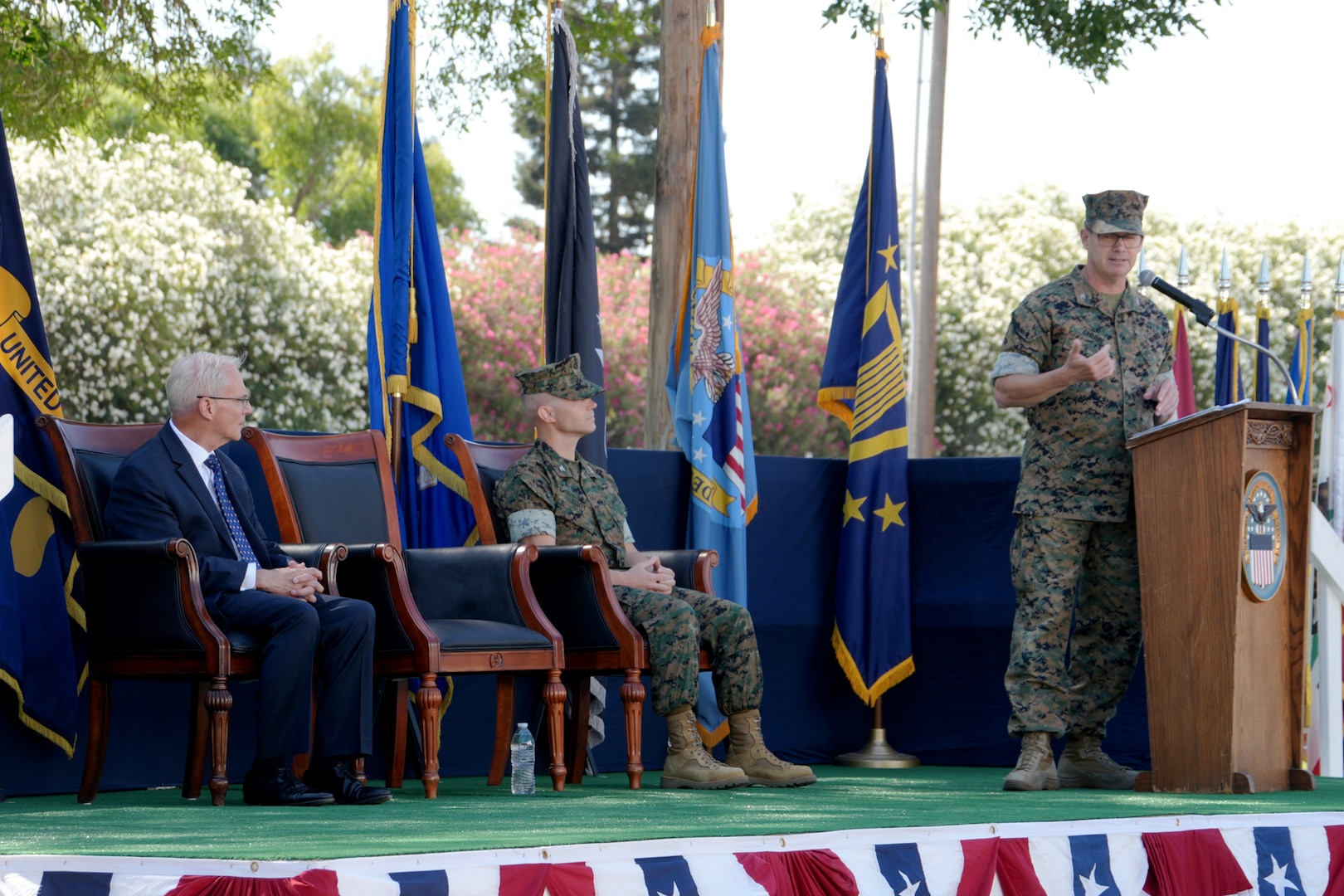 A man in a camouflage military uniform stands on a stage outdoors behind a lectern giving a speech.