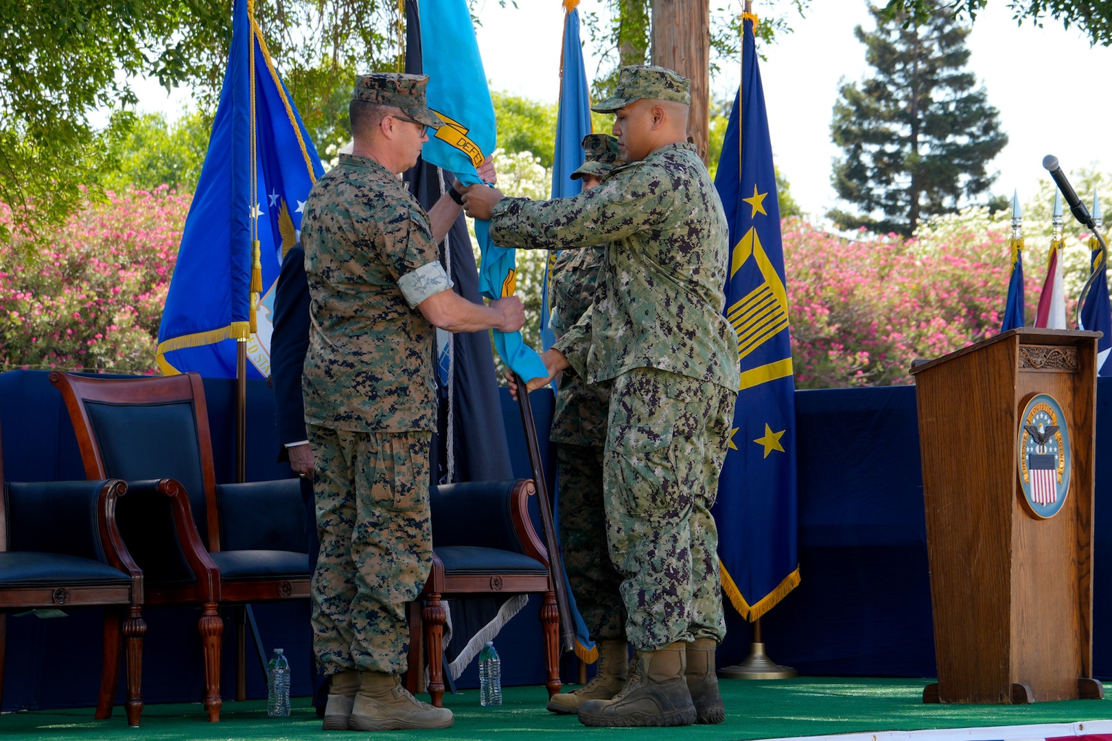 A man in a camouflage military uniform passes a blue flag to another man in a camouflage military uniform. There are other people in the background.