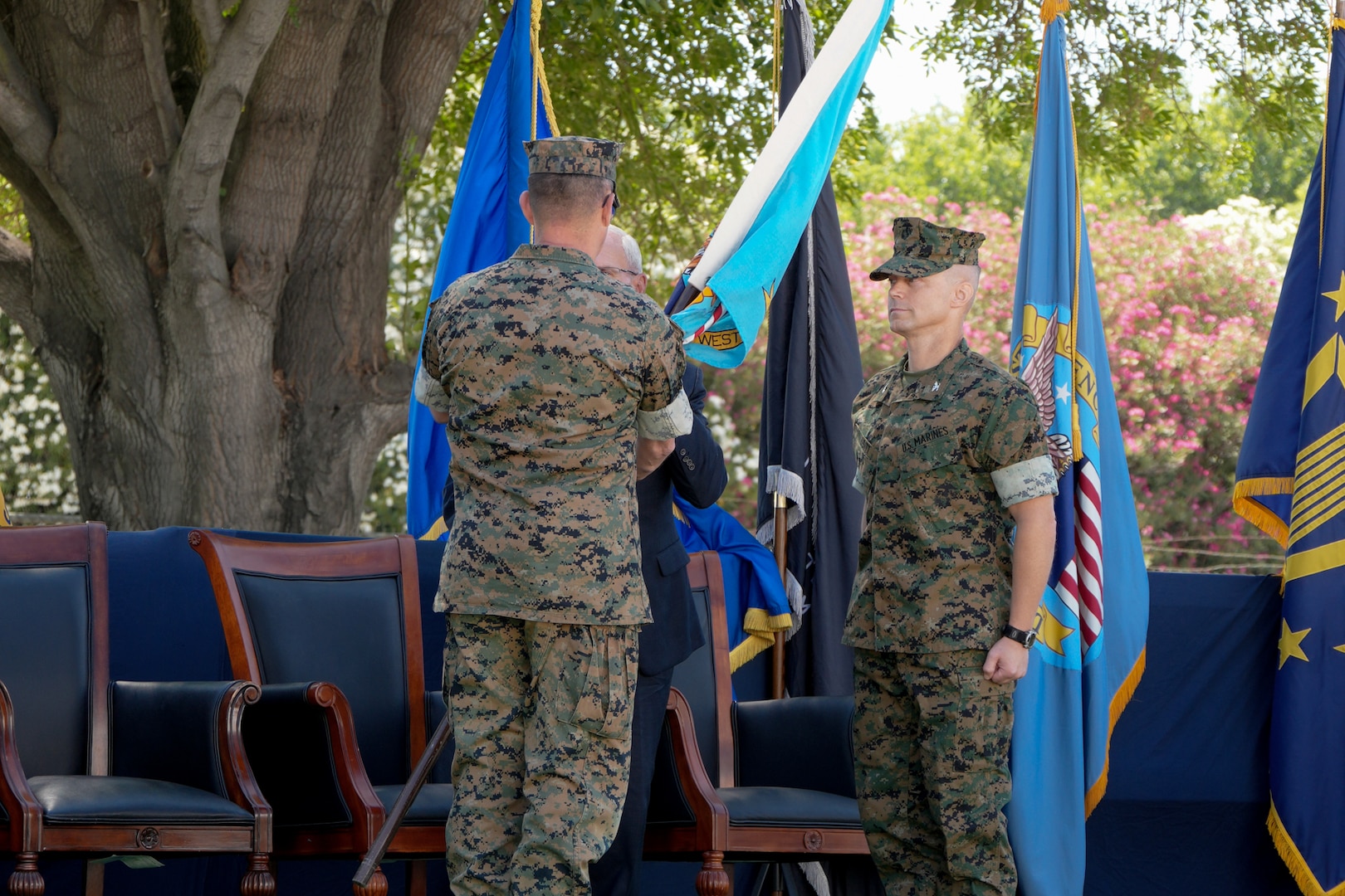 A man in a camouflage military uniform passes a blue flag to a man in a blue suit. There is another man in a camouflage military uniform standing to their right.