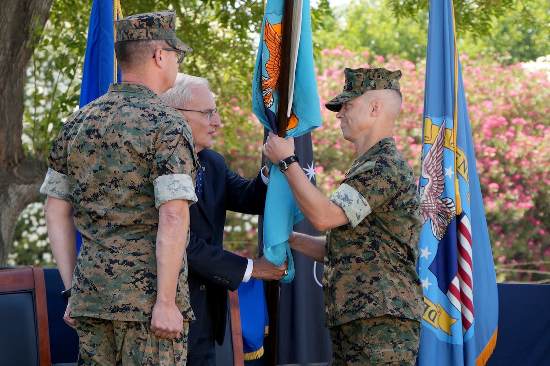 A man in a camouflage military uniform takes a blue flag from a man in a blue suit. There is another man in a camouflage military uniform standing to the left of them.