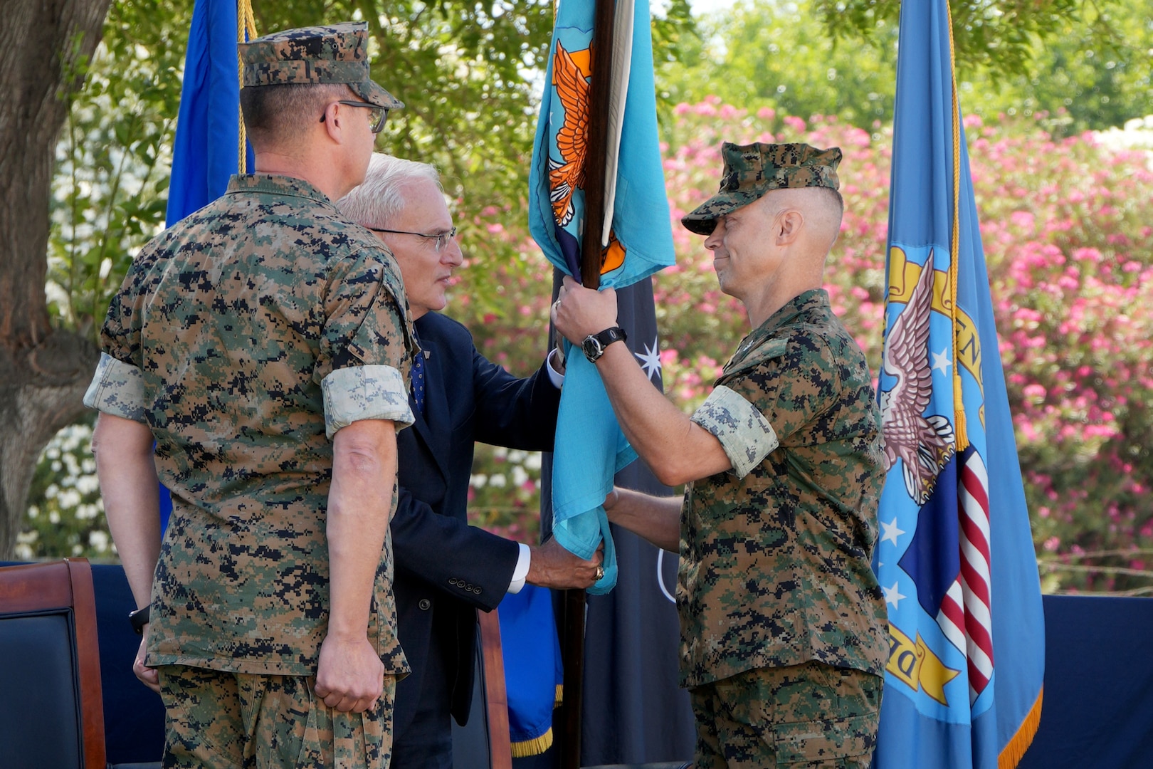 A man in a camouflage military uniform takes a blue flag from a man in a blue suit. There is another man in a camouflage military uniform standing to the left of them.