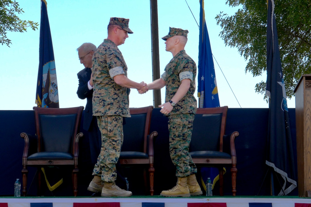 Two men wearing camouflage military uniforms stand on a stage outdoors shaking hands.