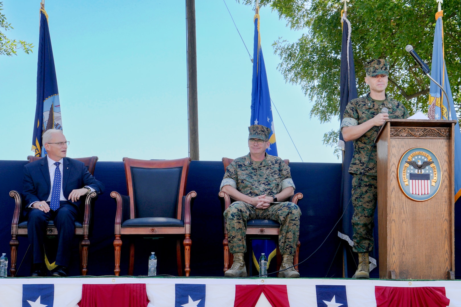 A man in a camouflage military uniform stands on an outdoor stage behind a lectern giving a speech.