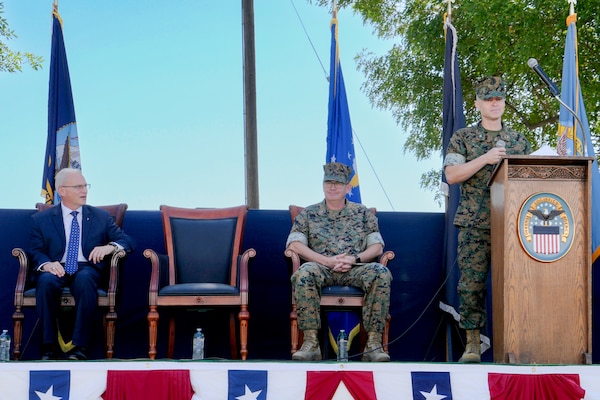 A man in a camouflage military uniform stands on an outdoor stage behind a lectern giving a speech.
