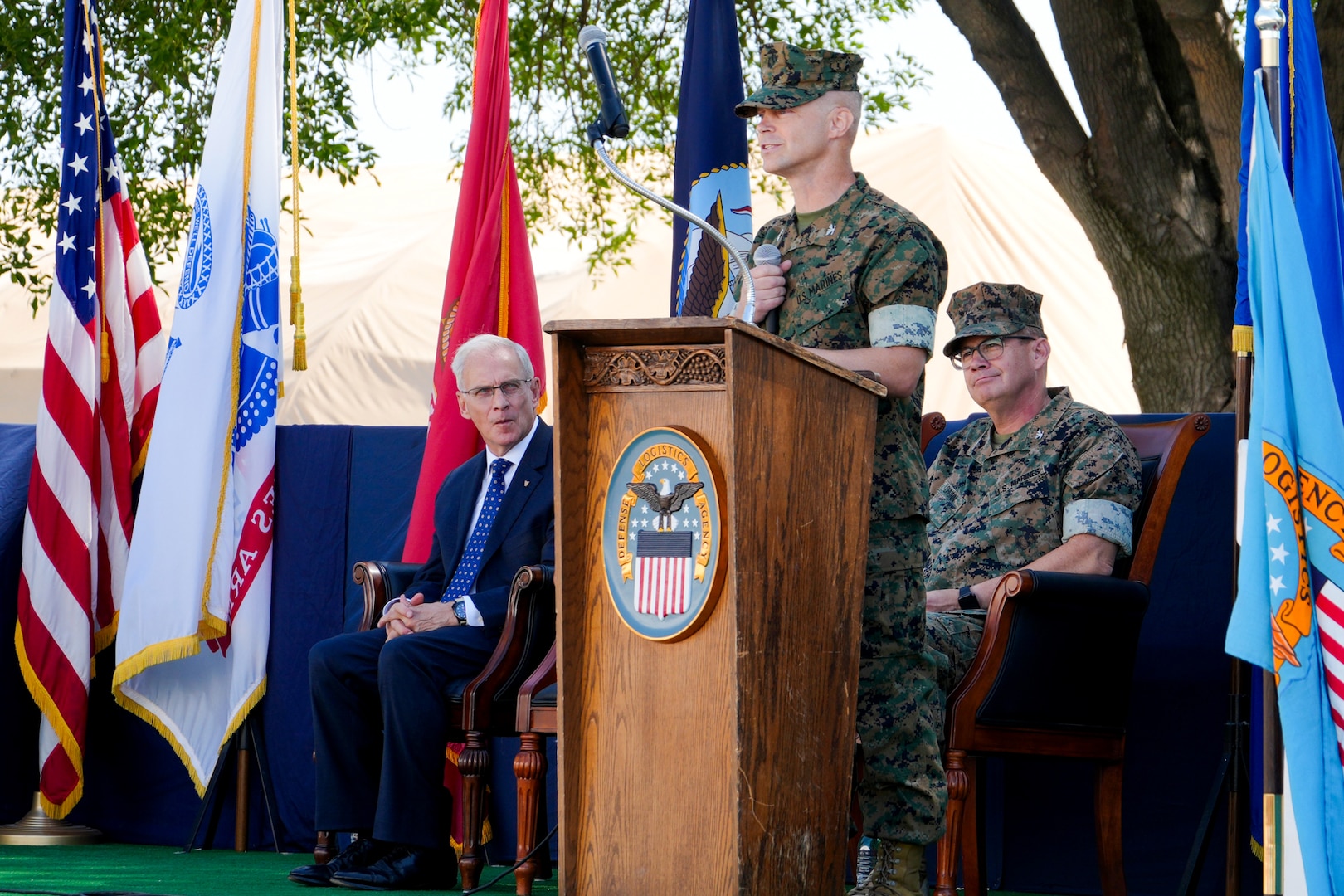 A man in a camouflage military uniform stands on an outdoor stage behind a lectern giving a speech.