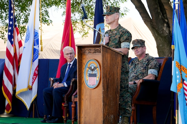 A man in a camouflage military uniform stands on an outdoor stage behind a lectern giving a speech.