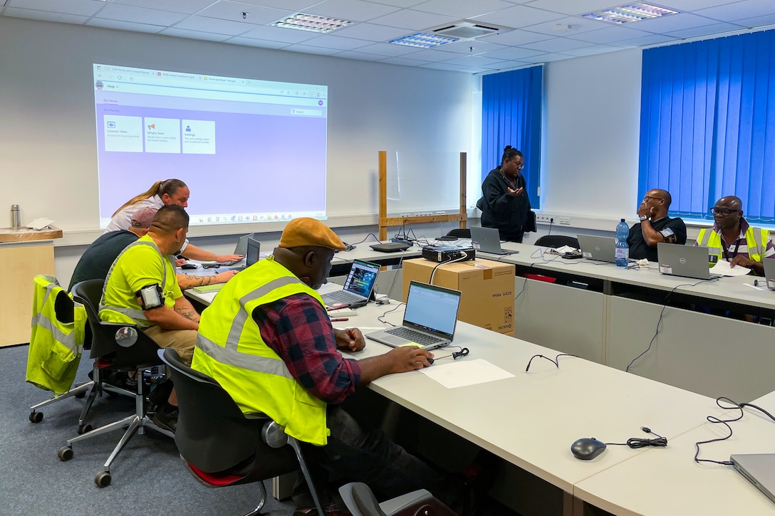 A group of people wearing reflective safety vests sit around a conference table working on laptop computers.