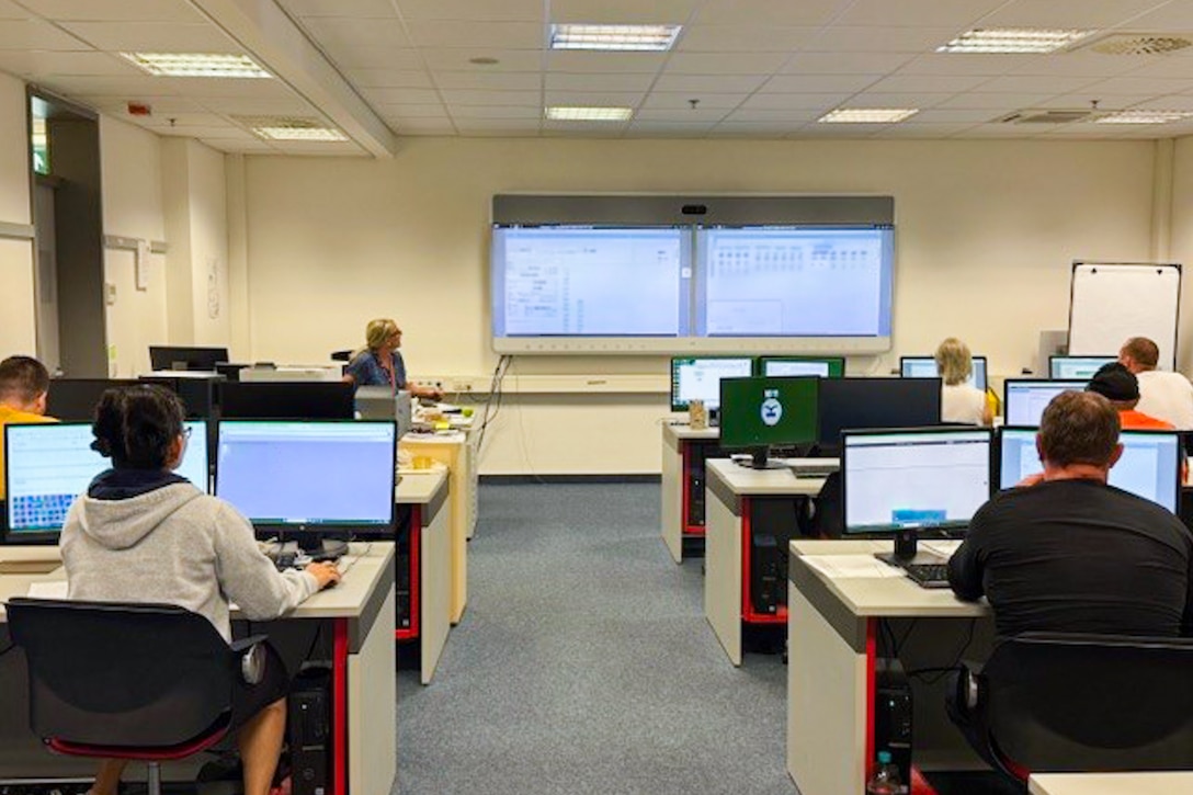 A group of people sit behind computers at desks facing a large projection screen on the wall in front of them.