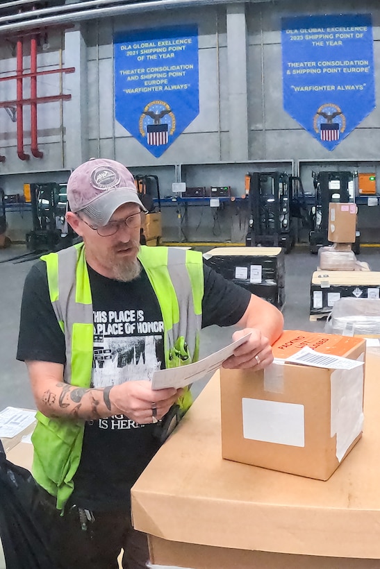 A man wearing a reflective safety vest looks at a piece of paper while resting his arm on a brown box next to him in a warehouse.