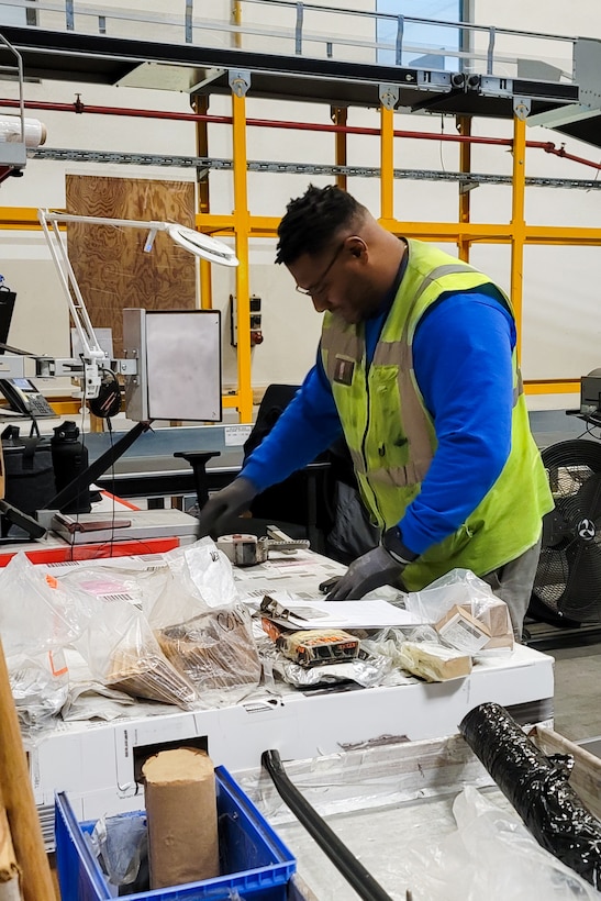 A man in a reflective safety vest looks through shipping labels and receipts at a table in a warehouse.